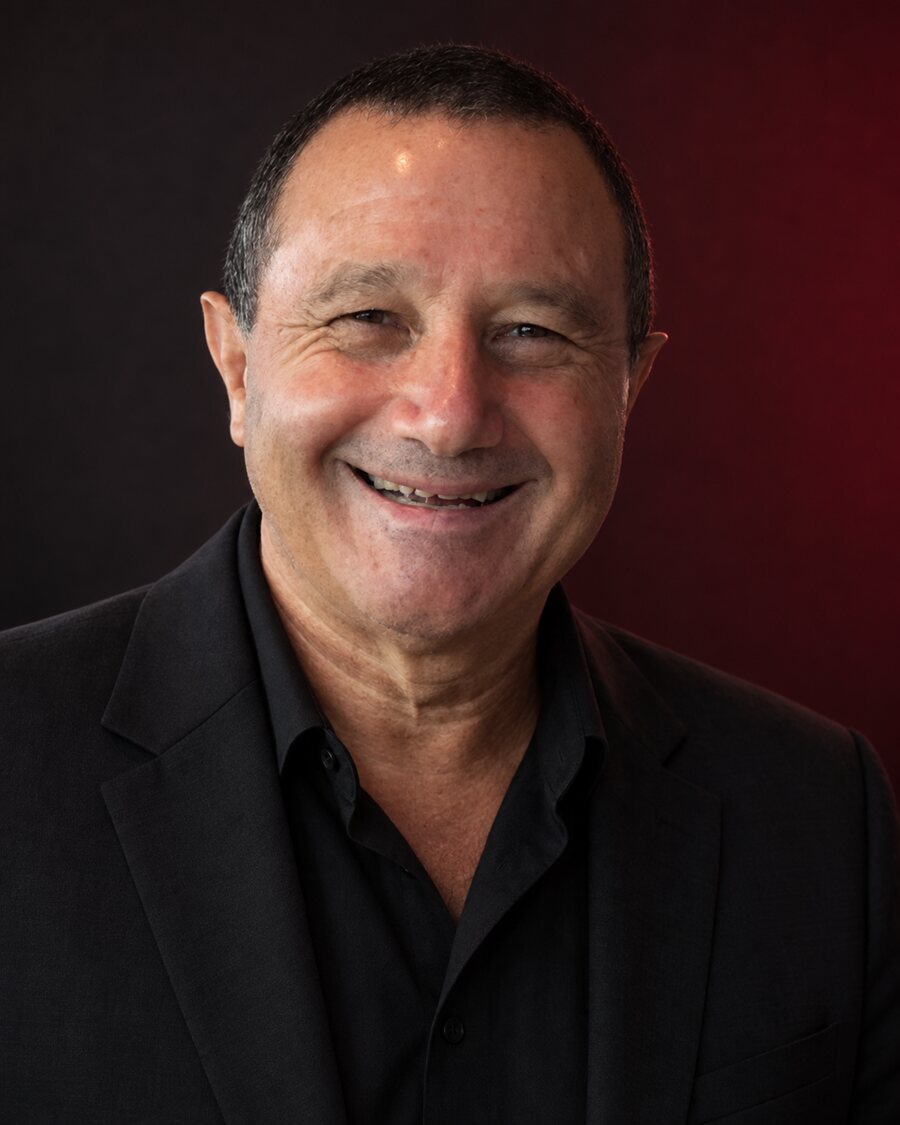 Barry, smiling warmly in a black blazer and open-collar shirt against a dark studio backdrop.